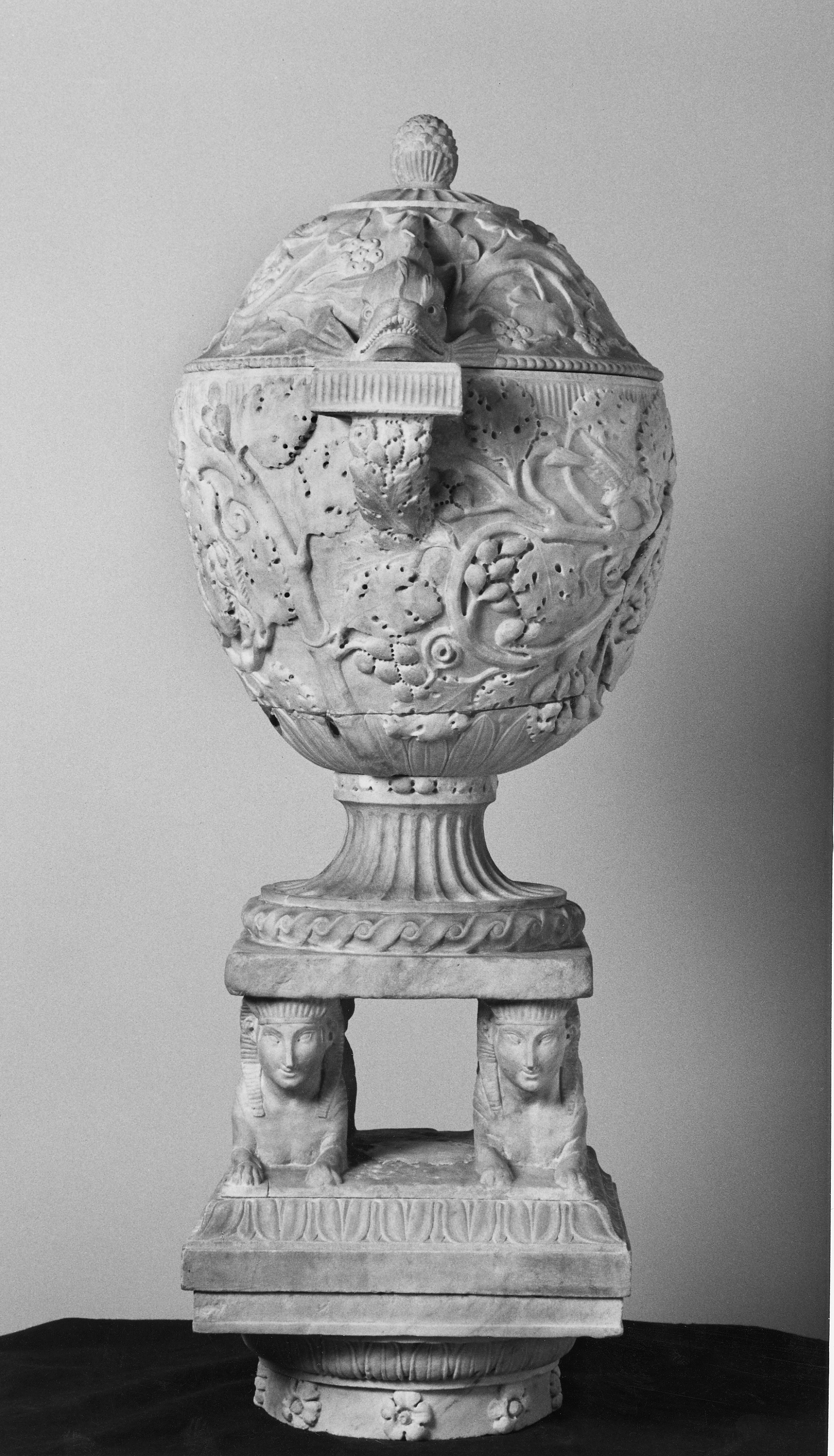 Schwarz-weiß Fotografie einer Vase (oder Aschenurne) aus Marmor im Profil. Die Mischwesen des Sockels sind frontal zu sehen.