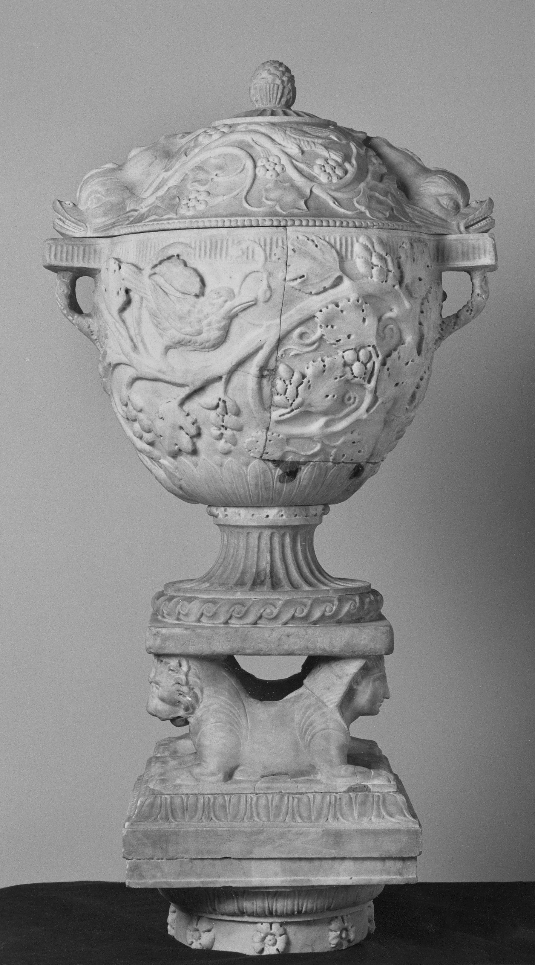 Schwarz-weiß Fotografie einer Vase aus Marmor, die auf einen Sockel mit Mischwesen montiert ist. 